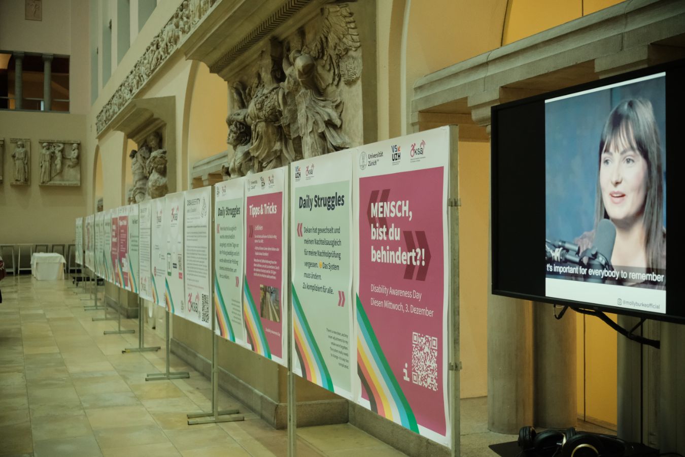 Plakatausstellung: Eine Reihe von Informationsplakaten zum Disability Awareness Day steht im Lichthof der UZH, daneben ein Bildschirm mit einem Video und Untertiteln.